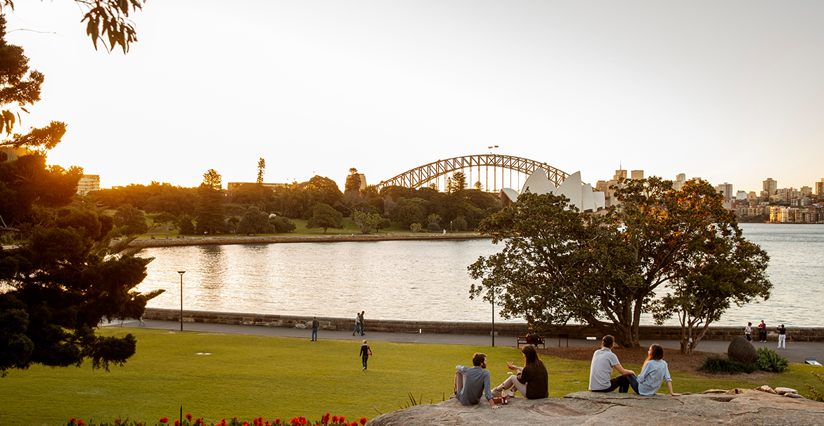 People relaxing on lawn overlooking Sydney Harbour Bridge, at sunset.