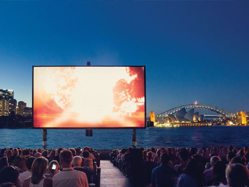 A seated crowd watch a big screen with the harbour and Sydney Harbour Bridge in the background