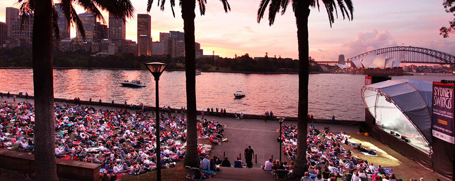 A crowd watching a concert in the Domain at sunset with harbour and harbour bridge in the background.