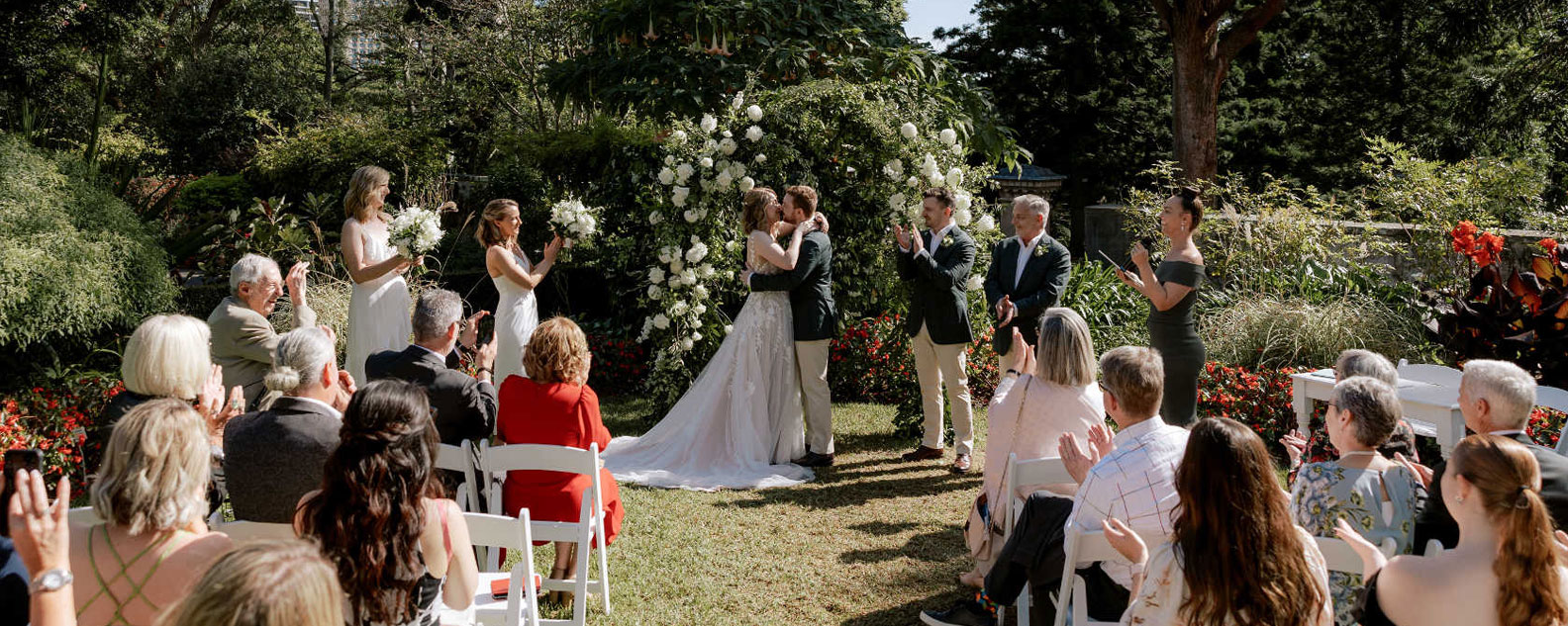 Couple kissing at a wedding ceremony with rows of seated onlookers in a garden setting.