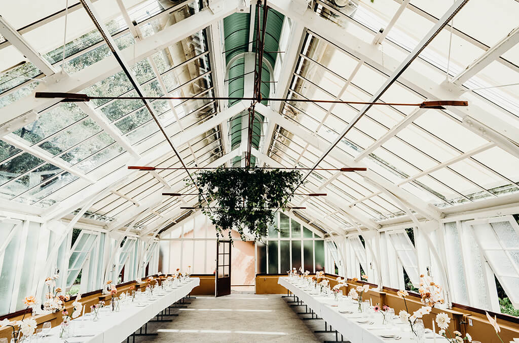 Interior of green house with glass roof and walls, and white tables set for a wedding