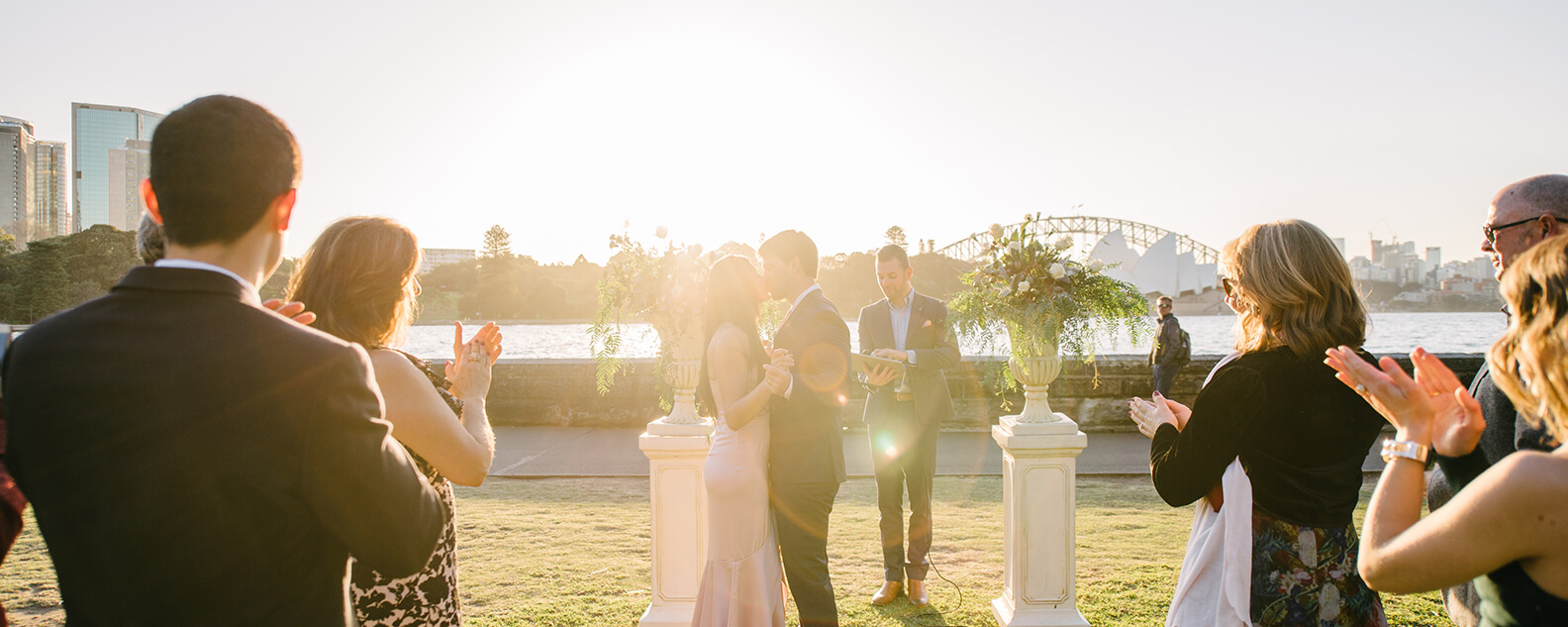 Bride and groom kiss at a wedding ceremony, with sun setting over Sydney Harbour behind them