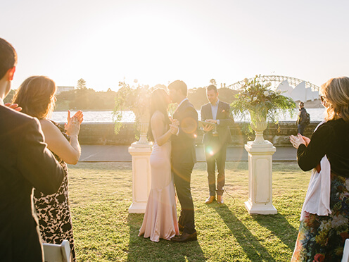 Bride and groom kiss at a wedding ceremony, with sun setting over Sydney Harbour behind them