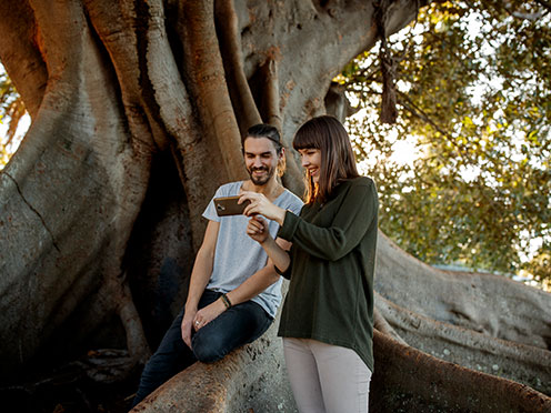 Couple resting near a large tree and smiling while looking at a phone screen