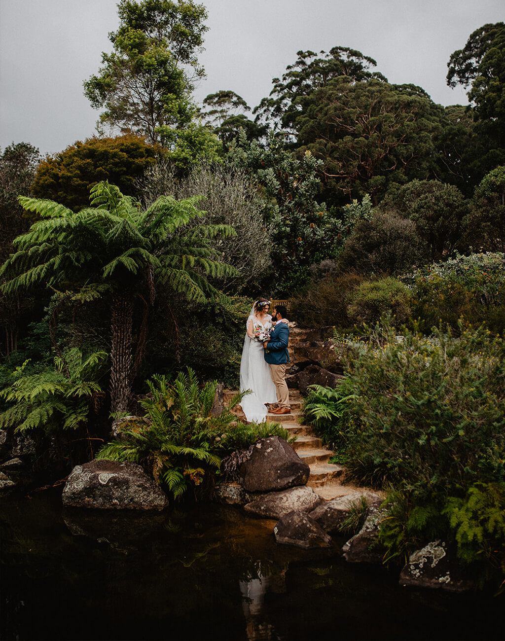 Bride and groom next to pond in lush garden with towering ferns and prehistoric-looking jungle