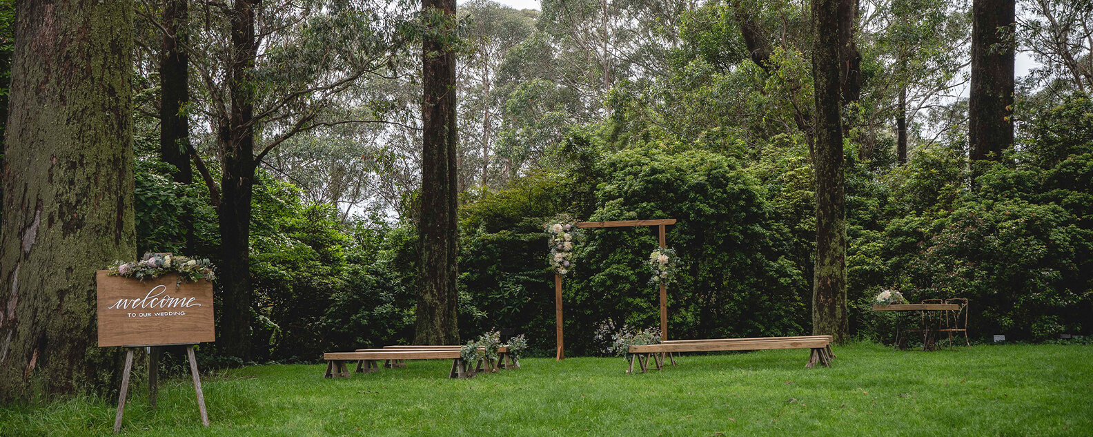 Lawn surrounded by tall eucalyptus trees, with rustic benches, flower arbour and a welcome sign ready for a wedding