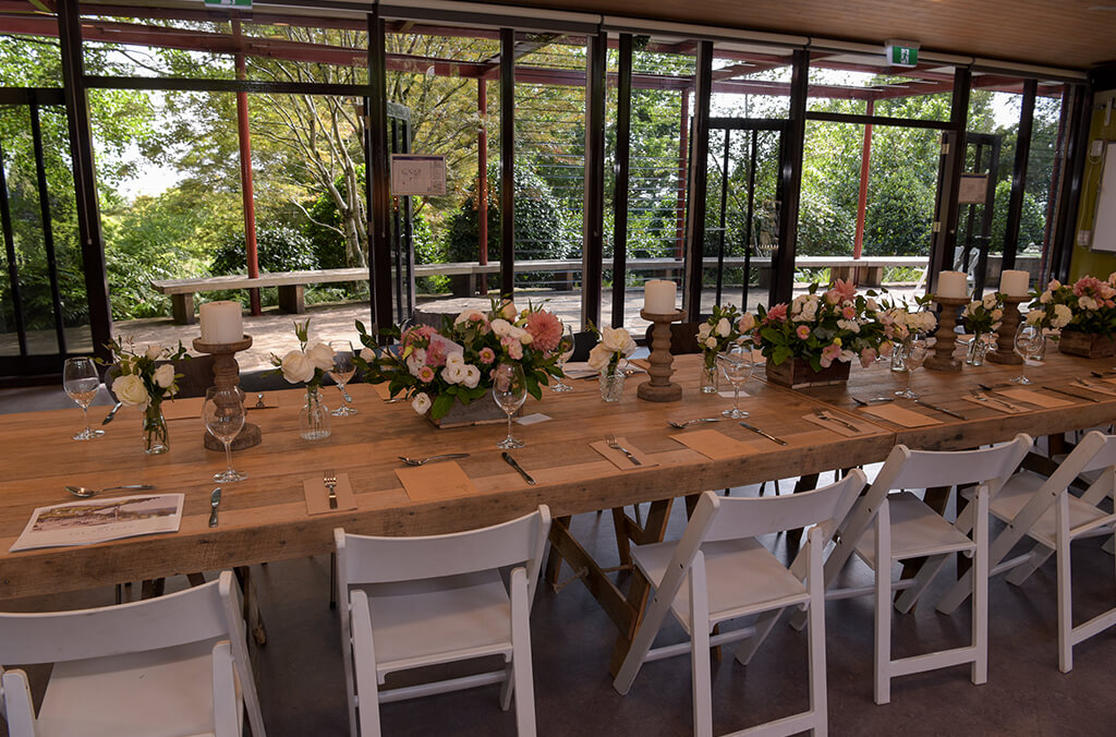 Room with floor to ceiling windows overlooking garden. Long tables set up for a wedding