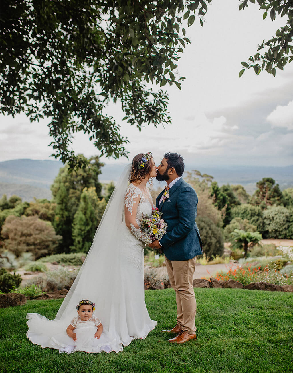 Bride and groom kiss overlooking views of Blue Mountains. Baby sits on bride's skirt
