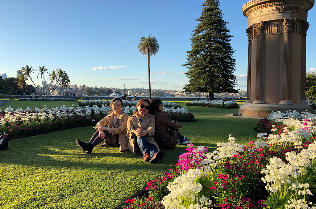 Group of friends sit on lawn smiling with colourful flower beds around them