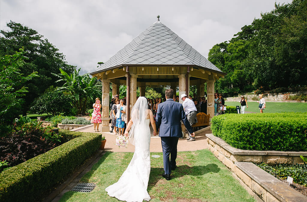 Bride and father walking to a sandstone pavilion through manicured garden