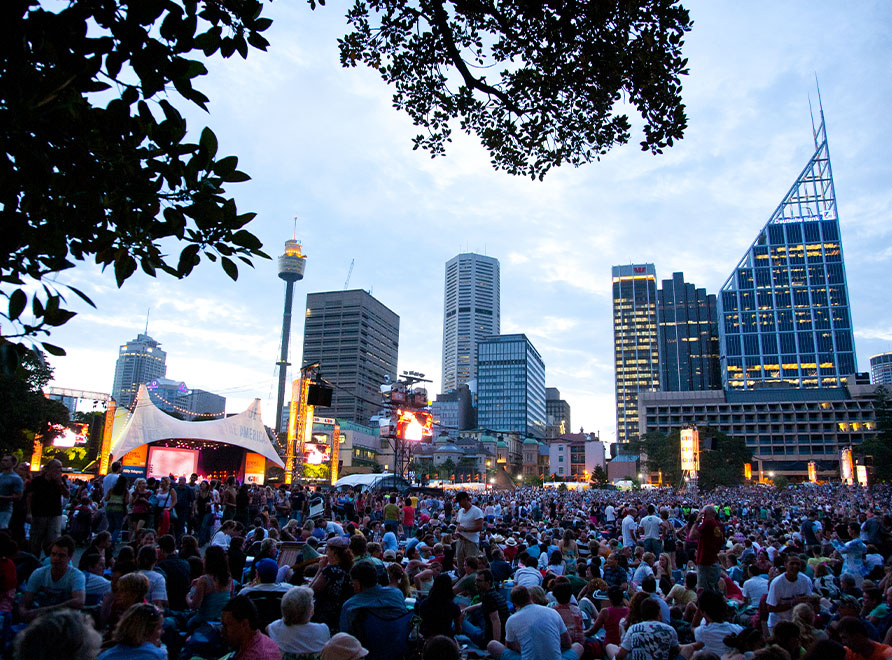 A large crowd attending a concert in The Domain with the cityscape in the background at dusk