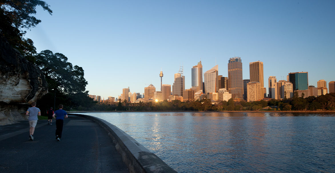 Three people running along The Domain running trail at sunset with a view of the water and city skyline on the right