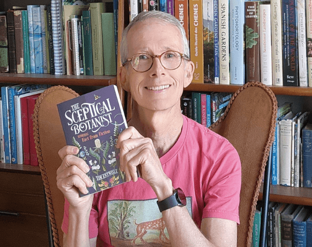 Tim Entwisle sitting in a brown leather chair, holding his book the Sceptical Botanist against a bookcase in the background