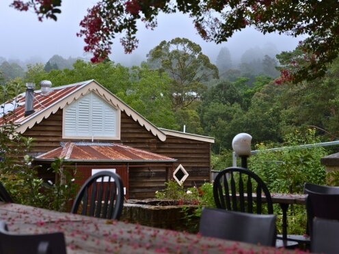 An old farmhouse with tables and chairs outside, misty mountains