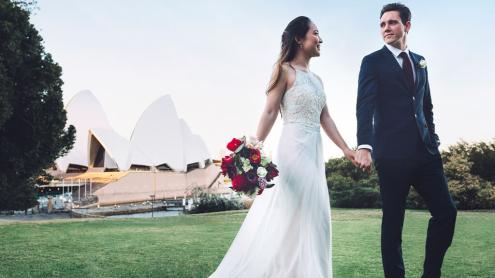 Bride and groom walk across lawn holding hands, the Sydney Opera House behind them