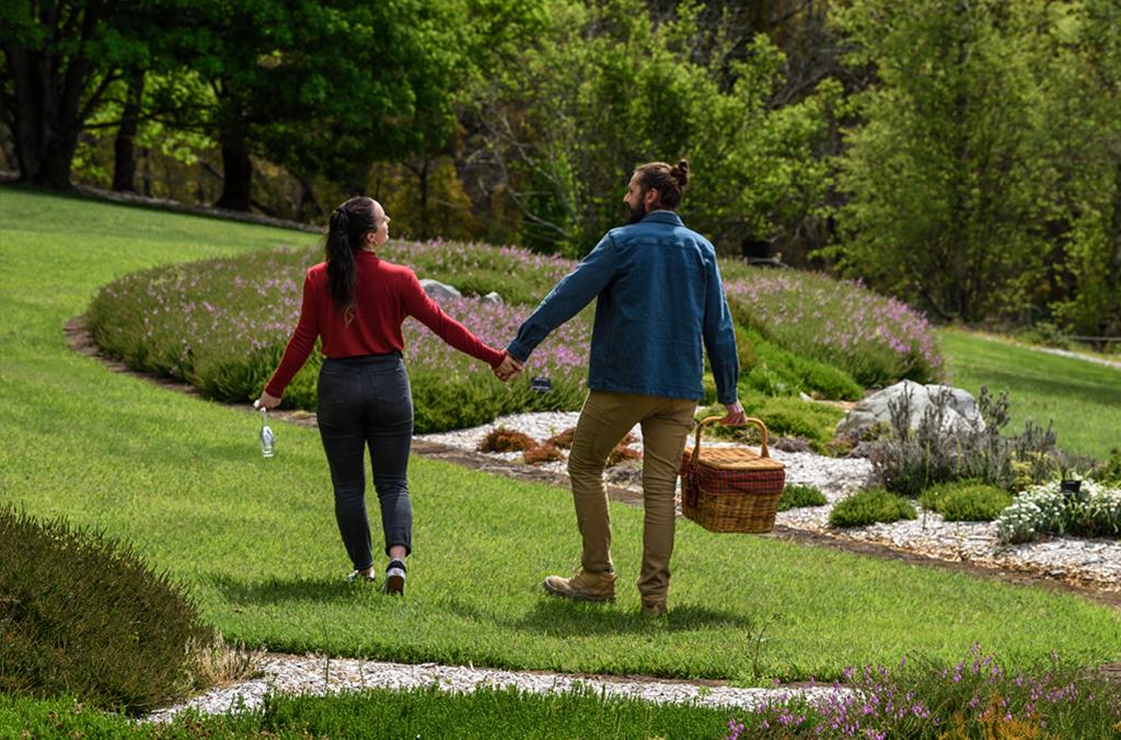 Two people hold hands, walking through heath garden, with a picnic basket