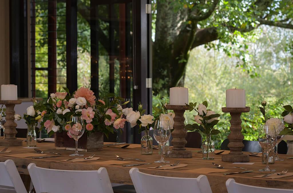 Open folded doors leading to a shady garden, a dining table set with wine glasses, candles and flower centrepieces