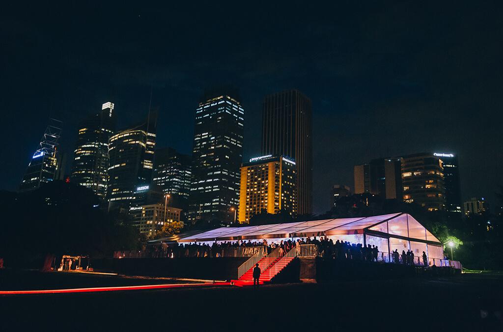 Marquee set up for an event on a lawn at night, with city skyline behind it