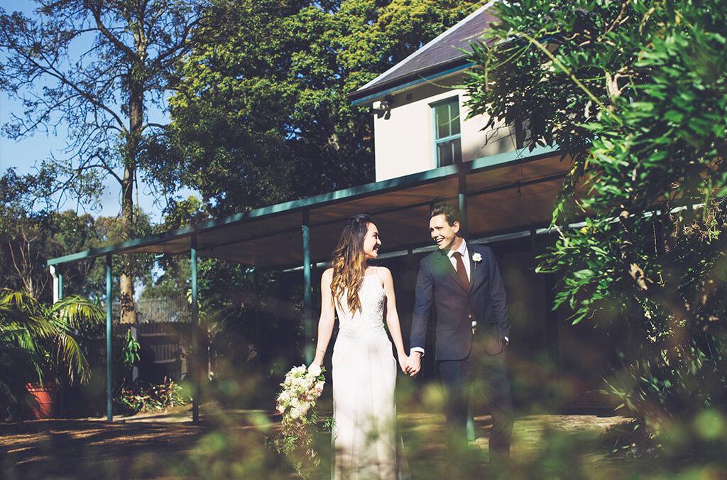 Bride and groom hold hands outside a charming, cream-coloured house