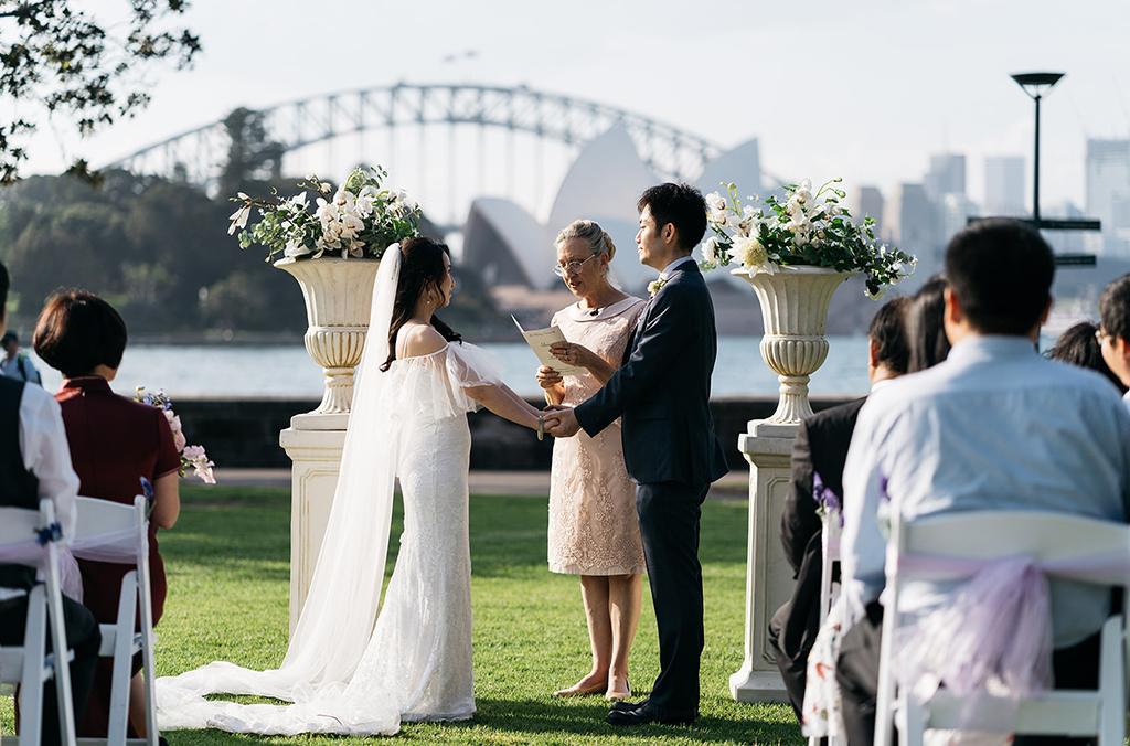 Bride and groom exchange wedding vows with celebrant on lawn overlooking Sydney Opera House