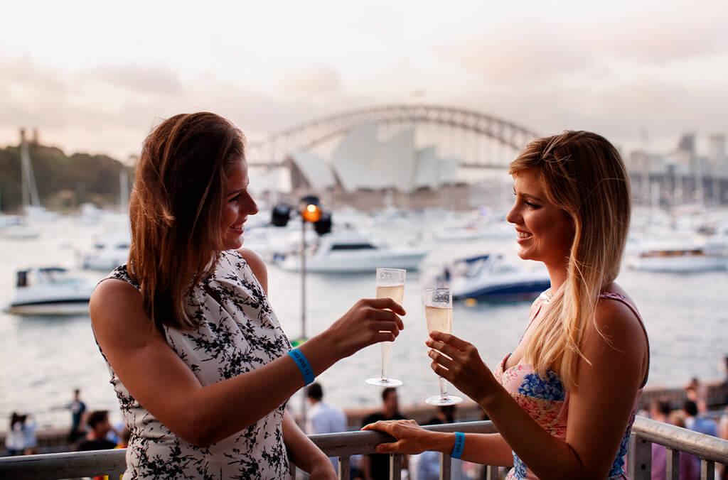 Two women cheers with sparkling wine, overlooking Sydney Harbour with Opera House and Bridge