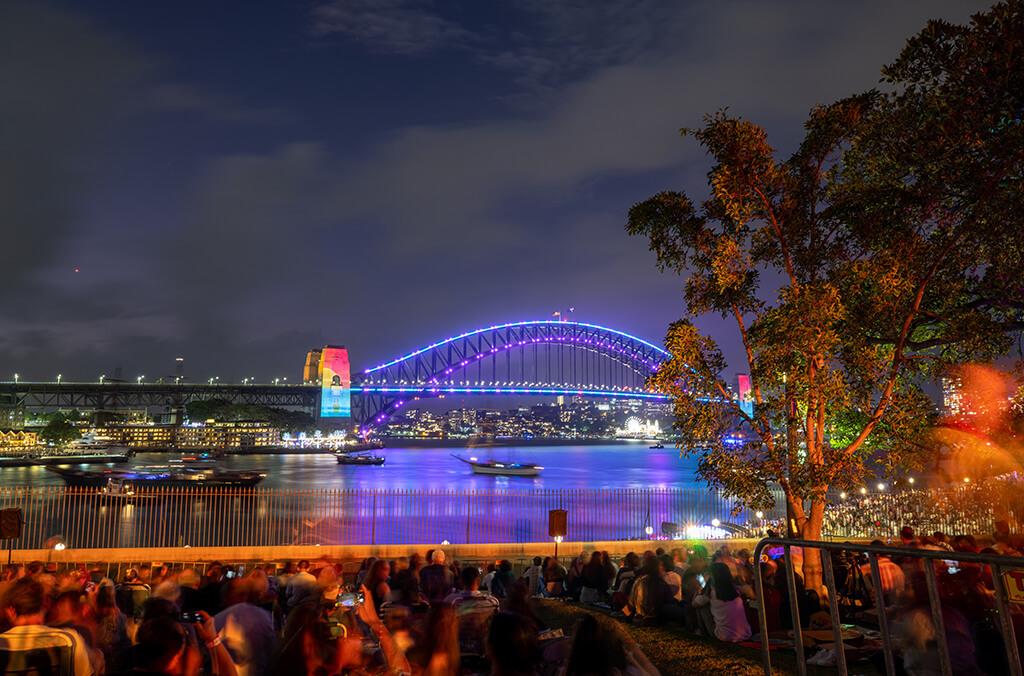 Crowd gathered at night on lawn overlooking Sydney Harbour Bridge lit up