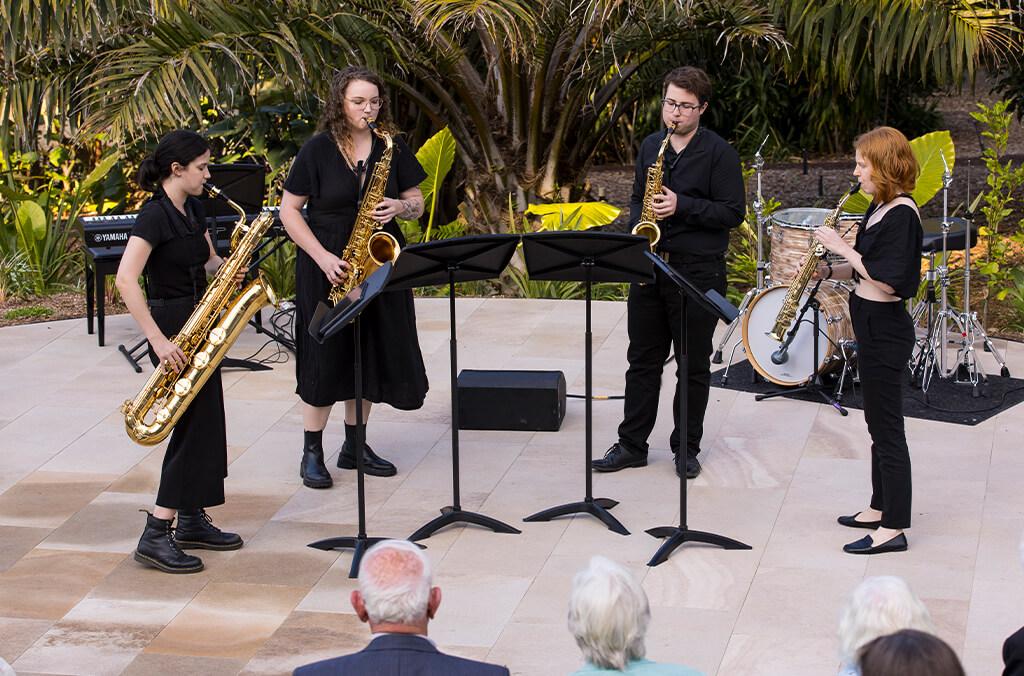 Four saxophonists perform in tranquil garden with seated crowd watching