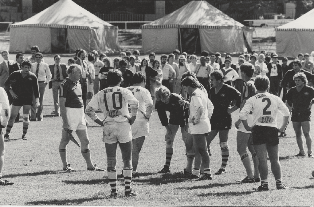 A group of footballers huddled during a game at the Domain, surrounded by spectators with marquees set up in the background, the Domain 1984