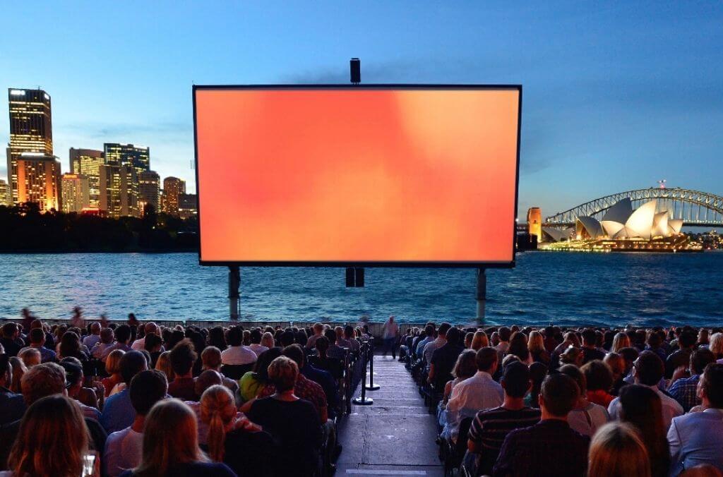 Giant film screen set up on Sydney Harbour at dusk, with crowd seated ready to watch a movie.
